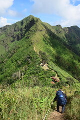 the extremely sharp grassy ridge leading to prominent peak with a small camp near the base of final camp and  the hiker walking with large backpack walking on the narrow ans steep path