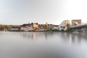 A long-exposure photograph capturing the imposing, Gothic architecture of a riverside church, complete with a tall spire, situated directly on the bank of a smooth, calm river, with a graveyard