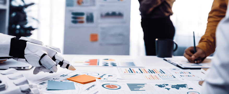 A futuristic business meeting showcasing a robotic hand analyzing charts and graphs, highlighting the integration of technology and team collaboration in a modern workspace.Noogenesis
