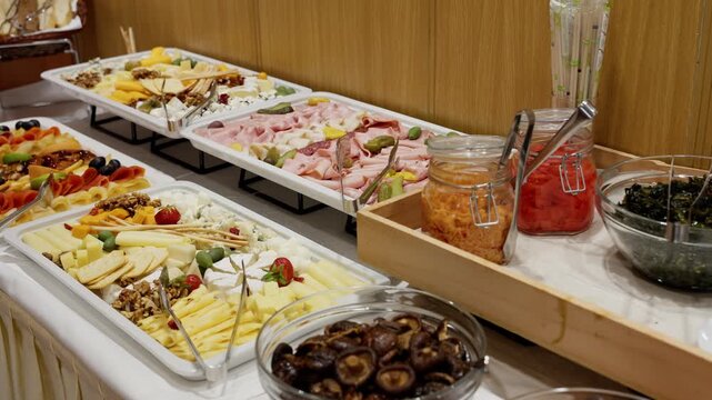Abundant cold buffet showcasing assorted cheeses, sliced cold cuts, pickles, olives, nuts, fresh fruits, and vibrant salads, arranged on elegant trays with tongs and jars of flavorful condiments
