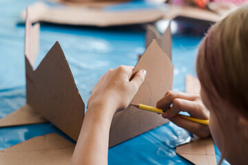 Tropical Christmas DIY art class with kids and diverse women workshop leaders making an eco-friendly cardboard holiday house, visible greenery and warm weather outside, boy wearing Santa hat proudly