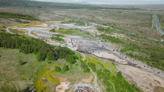 Aerial view of geothermal landscape near Strokkur Geyser. Drone shot capturing tourists exploring hot-spring pools and fumarole areas across the wide volcanic terrain under clear skies.