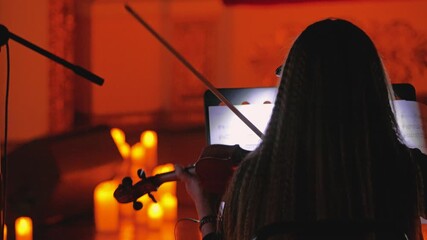 A female violinist plays by candlelight, her notes evoking the atmosphere of a chamber performance. Close-up, rear view