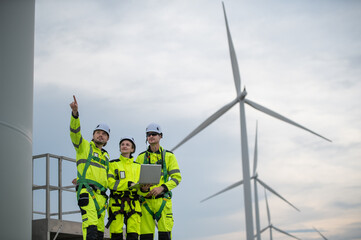 Engineers wearing safety helmets inspecting a wind turbine at a wind farm and holding a laptop while working.