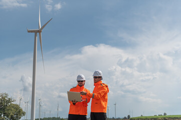 Engineers wearing safety helmets inspecting a wind turbine at a wind farm and holding a laptop while working.