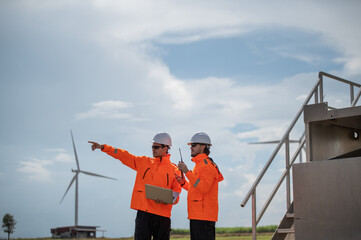 Engineers wearing safety helmets inspecting a wind turbine at a wind farm and holding a laptop while working.