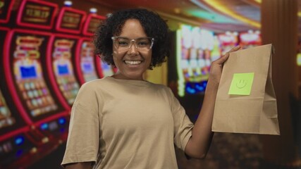 Young african american woman smiles and holds brown paper bag at slot machine in building; excitement.