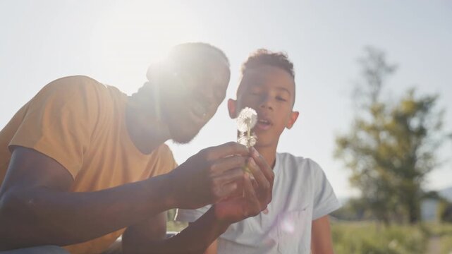 Smiling African American father and his son enjoying a sunny outdoors, making wishes and blowing dandelions, close up shot.