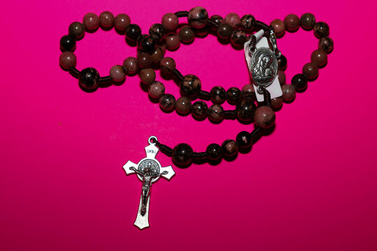 A rosary with mottled pink and brown rhodonite stone beads is laid out against a bright, saturated pink background. The metallic crucifix and the central medallion reflect a religious and spiritual th
