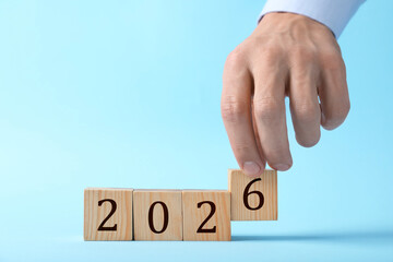 New 2026 year. Man putting cube with number 6 on light blue background, closeup