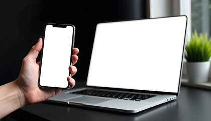 Hand holding smartphone with blank screen near laptop on a desk