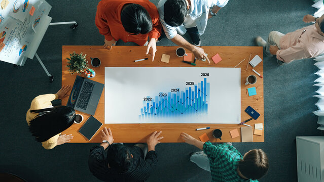 A diverse team of professionals collaborates over a table, analyzing a growth chart on paper. They use laptops and notes to discuss strategies in a modern office setup. Trope
