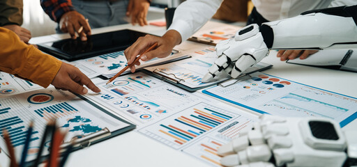 A dynamic workspace scene showcasing human hands and a robotic arm collaborating on detailed business reports and charts, emphasizing innovation and teamwork.Noogenesis