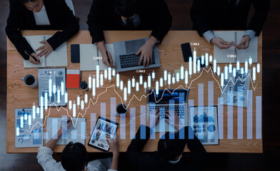 A group of professionals engages in a financial analysis session, reviewing stock market trends with charts, laptops, and reports in a collaborative office environment. Scalp
