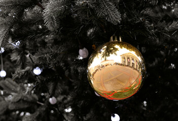 A golden Christmas ornament hangs from a dark pine branch, reflecting the festive atmosphere with its lights and architecture. Close-up. Selective focus.