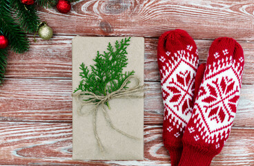Gift wrapped in craft paper with green branches, red knitted mittens on a rustic wooden background. Cozy Christmas flatlay composition with festive decorations. Top view. Close-up.