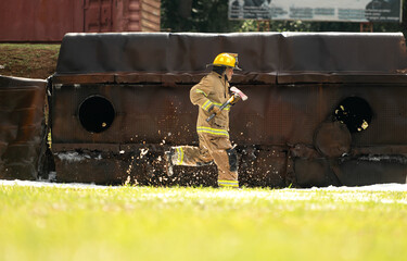 A firefighter in full turnout gear runs across a wet training ground while carrying a rescue axe