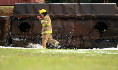 A firefighter in full turnout gear runs across a wet training ground while carrying a rescue axe