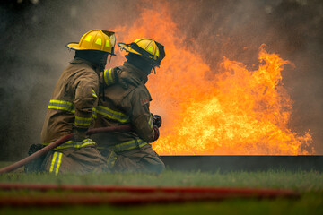 Naklejka premium Two firefighters kneel in position while directing a powerful water stream at a large blaze