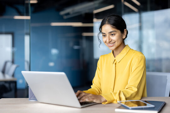A young Indian businesswoman in a yellow shirt sits at a desk in the office and works on a laptop with a smile - Powered by Adobe