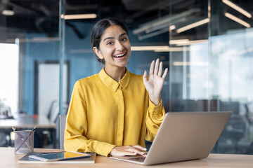 Portrait of a smiling young Indian woman sitting at an office desk, working on a laptop, greeting and waving at the camera