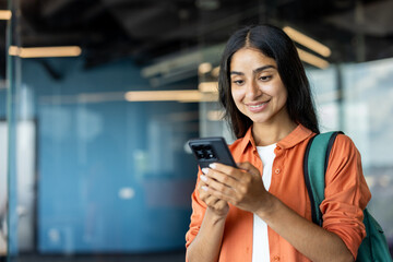 Young Indian smiling student girl in a red shirt and green backpack standing indoors and using a...