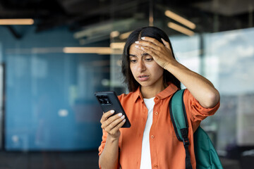 Close-up photo of a young Indian woman with a backpack and a shirt standing indoors and looking at her phone in shock, holding her head in disappointment