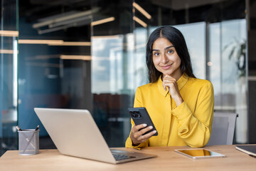 Portrait of a young smiling Indian woman in a yellow shirt sitting at an office desk and using a mobile phone