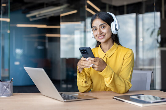 Portrait of a smiling Indian woman wearing headphones sitting in an office, using her phone and looking at the camera