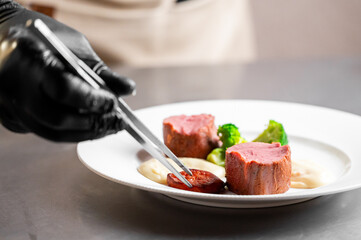 A chef in black gloves meticulously arranges seared beef filet mignon and steamed broccoli on a white plate with a creamy sauce using plating tweezers