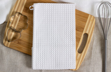 Mock-up of a white kitchen waffle towel on a wooden cutting board with steel cookie cutters and a whisk. Styled photo mockup for custom design