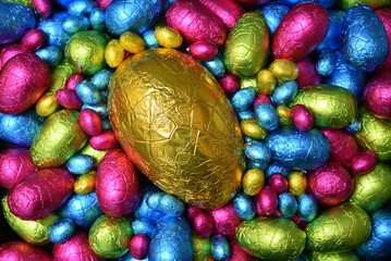 Pile of colourful foil wrapped chocolate easter eggs in pink, blue, yellow and lime green with a large gold egg in the middle.