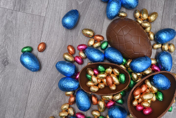 Pile  of colourful foil wrapped chocolate easter eggs in pink, blue, gold and green. Large halves of a brown milk chocolate egg have mini eggs inside, on a grey wooden background.