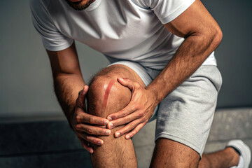 A man has a knee injury with a red scratch on his knee, holding the injured area with his hands