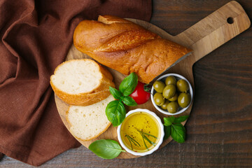 Tasty baguette served with oil, tomato, olives and basil on wooden table, top view