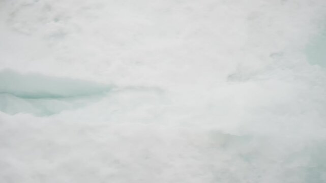 Fractured sea ice with visible turquoise meltwater seams and drifting floes transitions into soft, compacted slush and snow, revealing textures and patterns of a frozen seascape in seasonal thaw.