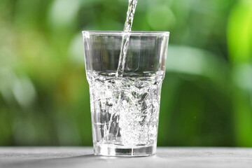 Pouring water into glass at grey table against blurred green background, closeup