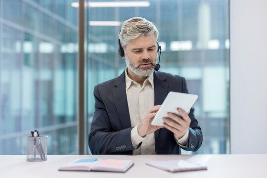 Professional mature businessman wearing a headset and working on a digital tablet in a modern office, providing customer service while looking at the screen