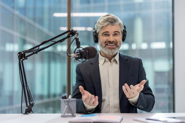 Mature businessman with gray hair and beard wearing headphones and suit jacket, smiling and gesturing while recording a professional podcast or radio show in a modern office studio