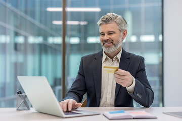 Happy mature businessman sitting at an office desk, smiling while holding a credit card and using a laptop for online shopping and secure e-commerce transactions