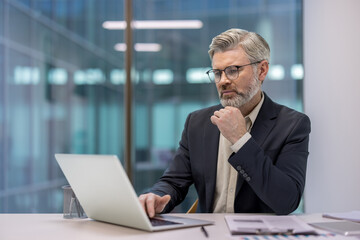 Thoughtful senior businessman working on a laptop in a modern office, planning business strategy...