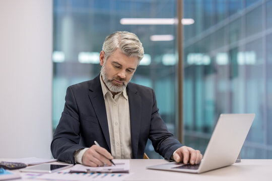 Mature businessman in suit studies financial reports and works on laptop at office desk, focused on charts, graphs and data while planning strategy in a modern corporate setting