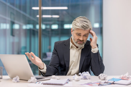 Businessman experiencing burnout while sitting at office desk, overwhelmed by work, with crumpled papers surrounding a laptop, reflecting frustration and difficulty in problem-solving