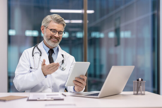 Male doctor in white coat and stethoscope smiles during a telemedicine video consultation, explaining treatment and offering remote care via tablet from a modern office setting - Powered by Adobe