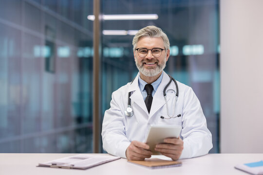 Mature male healthcare professional wearing a white lab coat and stethoscope, smiling while using a digital tablet at his office desk, symbolizing modern medicine and telehealth - Powered by Adobe