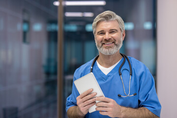 Confident senior male doctor in scrubs with stethoscope smiles while holding a tablet in a modern clinic hallway, conveying expertise, warmth and digital-ready patient care