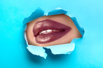 Young woman with beautiful lipstick, closeup. View on lips through torn hole in light blue paper