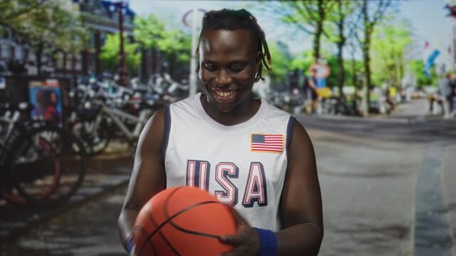 Man holding basketball on shoulder in street wearing usa white jersey with flag patch and blue wristbands, smiling and looking down at ball; team pride.