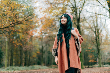 Young Woman wrapped in warm Shawl Poncho enjoying autumn walk in forest with fall yellow leaves. Travel lifestyle. Feeling harmony, reunion with nature. Relaxing, mental recovery. Local travel.