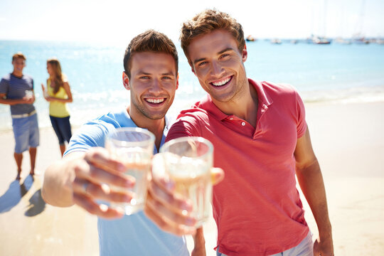 Two Caucasian young men raise glasses on a sunlit beach to celebrate together.